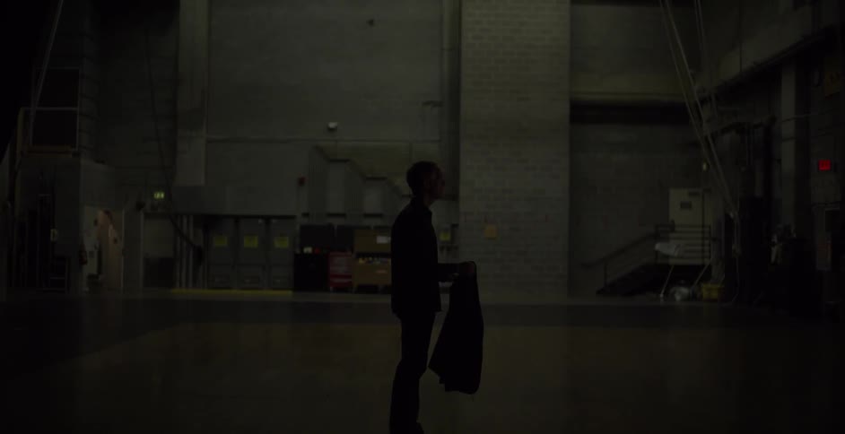 A man stands in the middle of a dark room removing his jacket