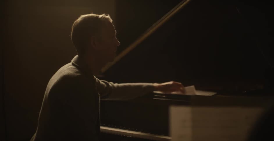 A man speaks in front of a piano then moves some papers