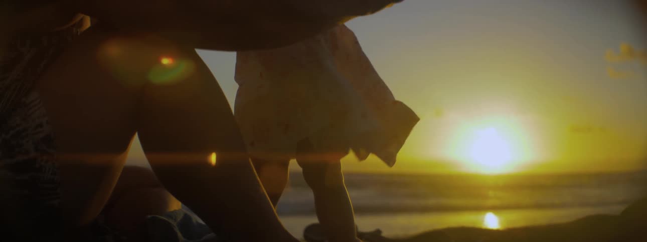 A mother helps her baby daughter stand as they sit on a beach at sunset