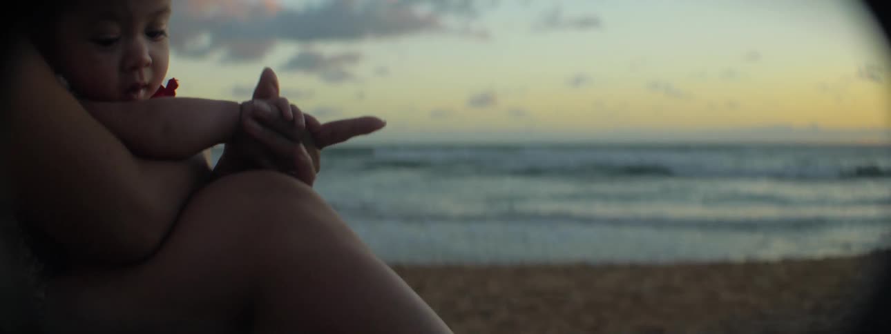 A woman plays with her baby's hand as they sit on the beach at dusk