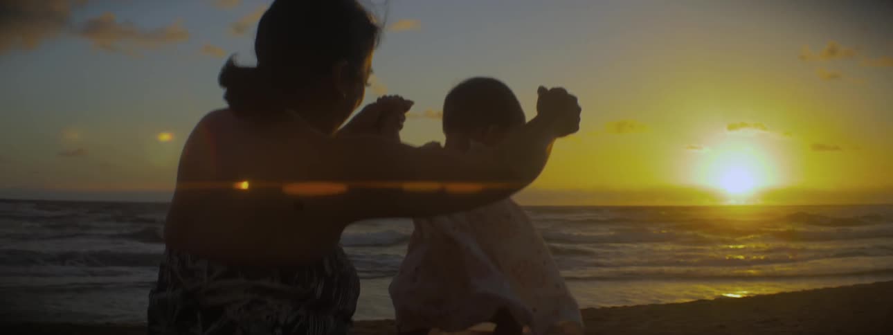 A woman helps a toddler stand in the sand at sunset
