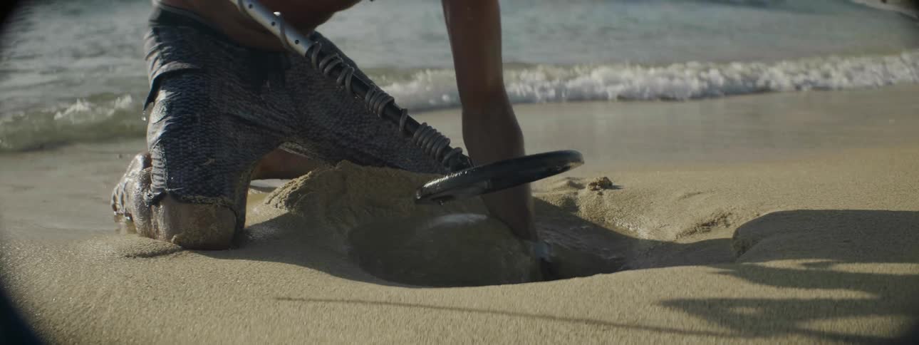 A man holding a metal detector digs through wet sand