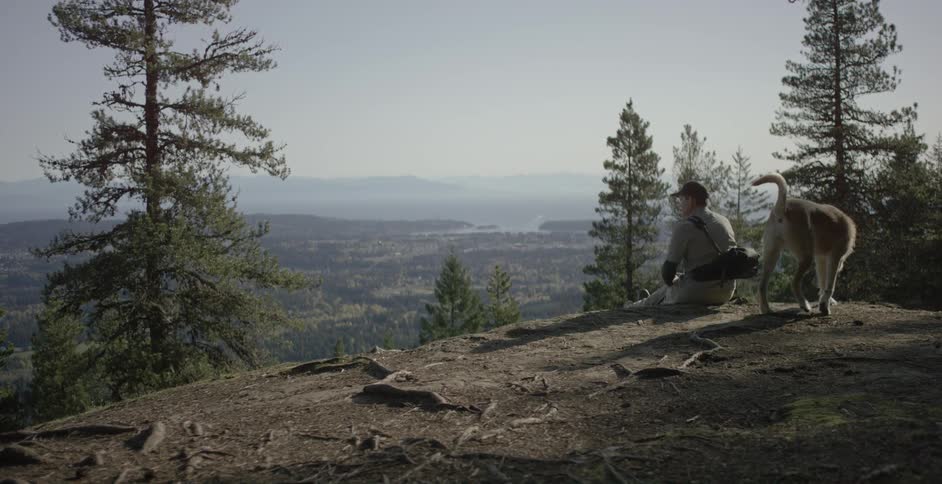 A man sits with his dog sits and looks out at a valley below him