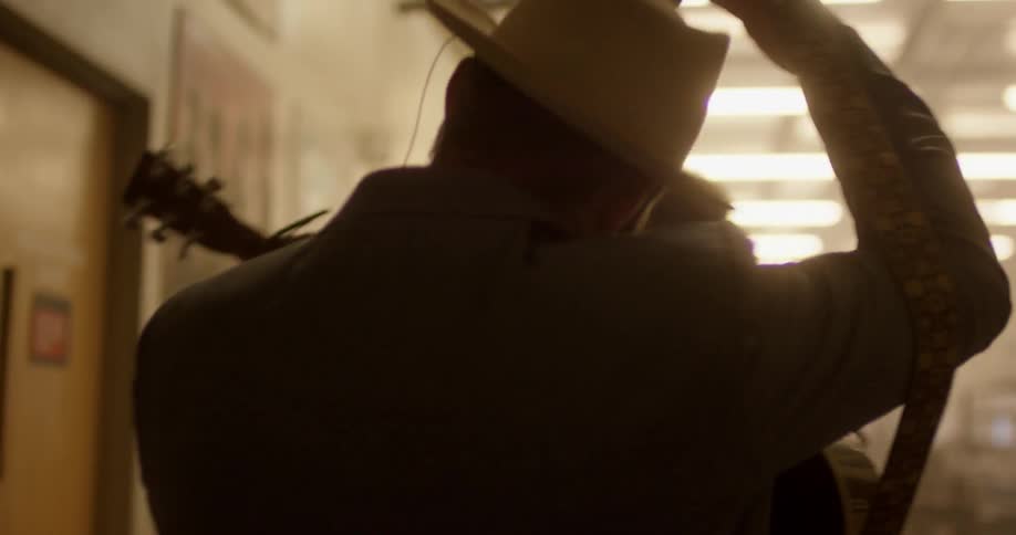 A man is led down a hall holding a guitar