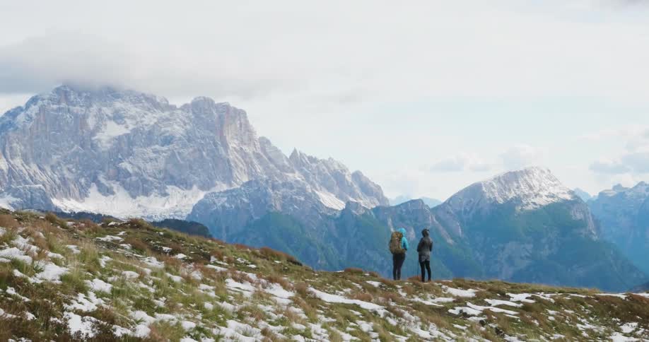 Two women stand on a mountain ridge looking at a snow-covered mountain range