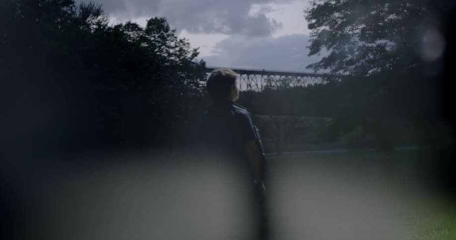A boy is stood still looking at a body of water near a train bridge