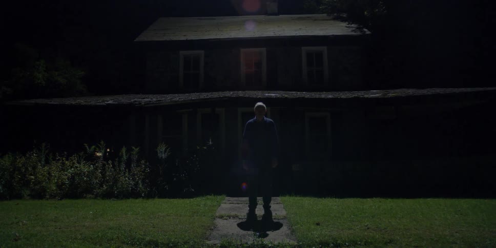 An old man stands in front of a rundown home at night
