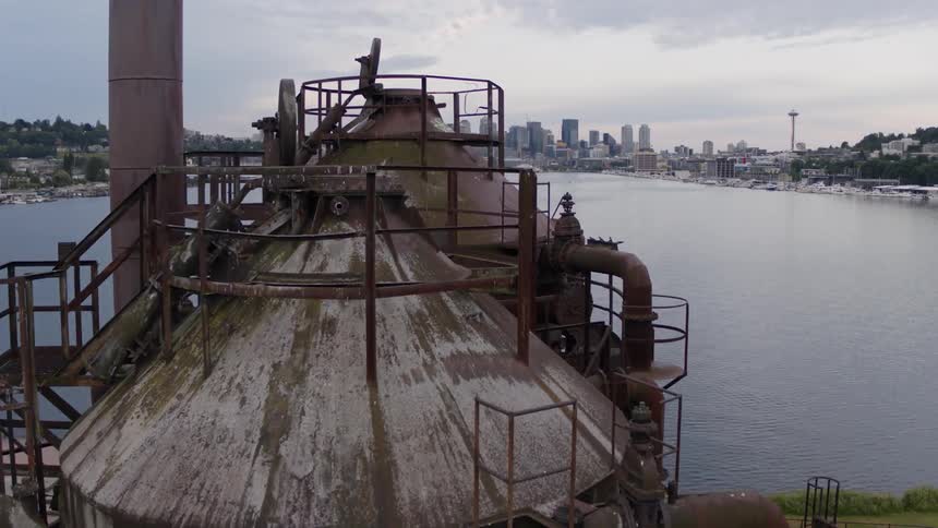 Skimming over an old abandoned gas works plant along a lake in front of a city skyline