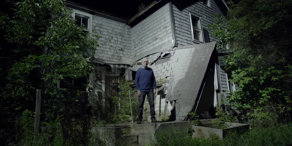 An older man stands near a broken house