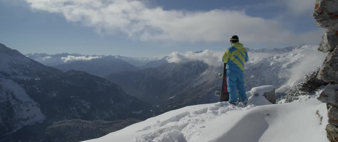 A man stands atop a snowy mountain with a snowboard
