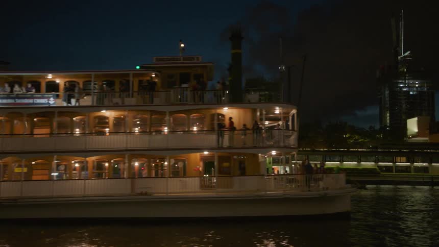A lit up boat on a river next to a city at night