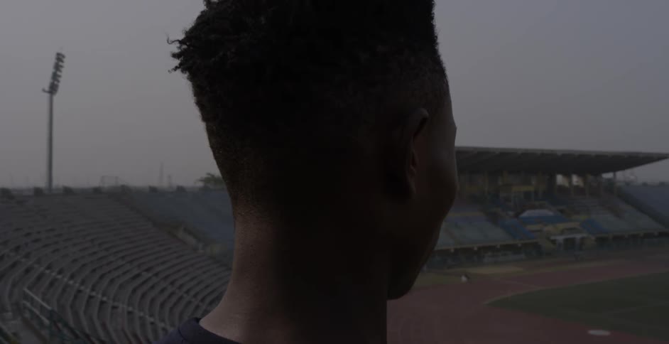 A young black man stands high up in the bleachers in a stadium looking down