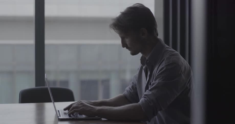Man sitting at a table next to a window typing on a laptop