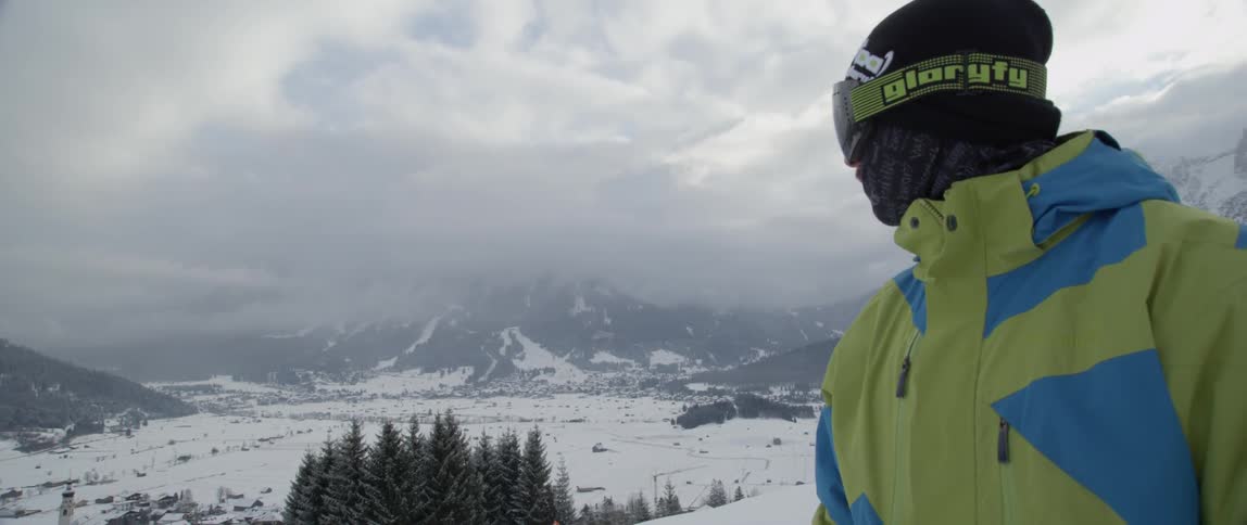 A man wearing snowboarding gear is looking backwards toward a mountain
