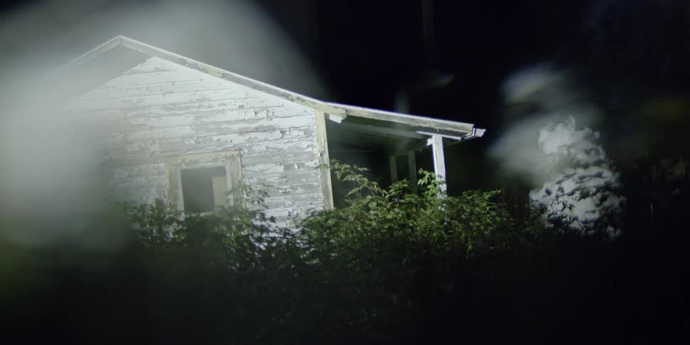 Headlights shine on an old house surrounded by bushes