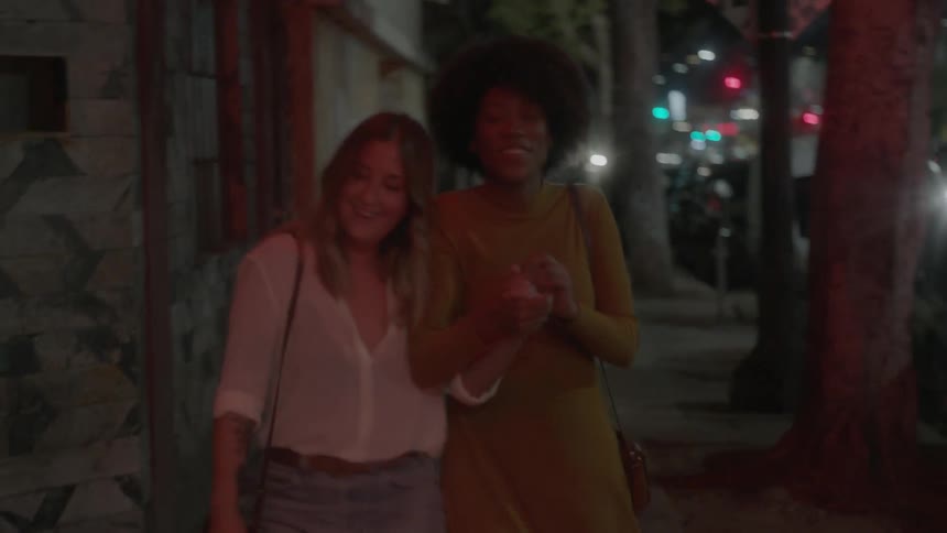 Two women exit a building holding hands and laughing at night