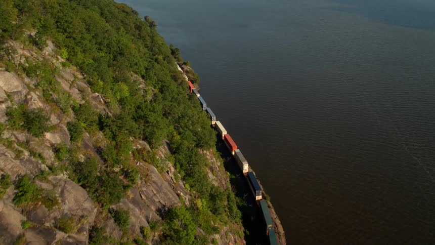 A train travels between a tree covered mountain and a lake