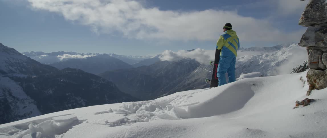 A snowboarder stands on top of a mountain looking at the scenery around him