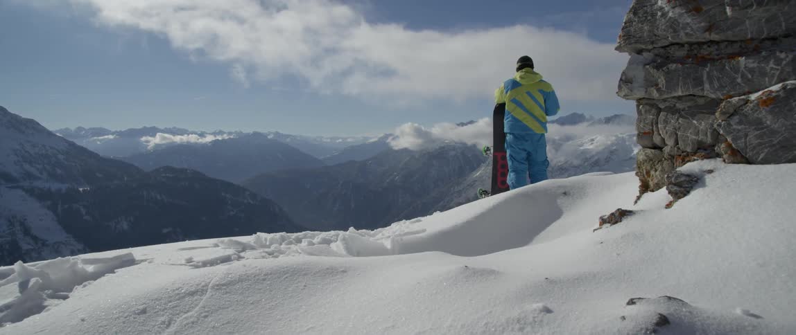 A man with a snowboard stands on a mountain peak taking in the view