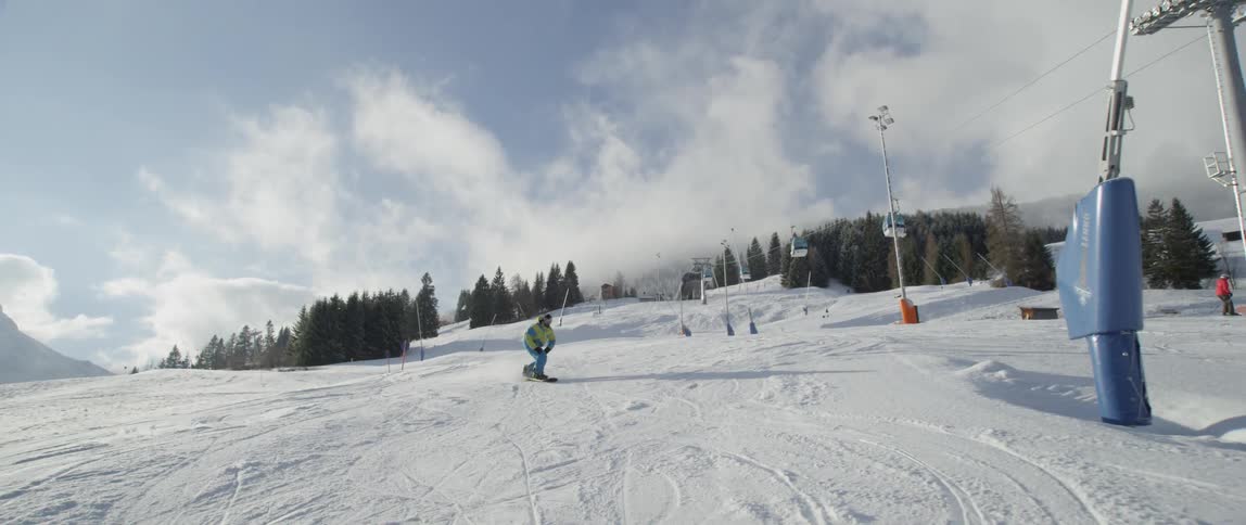 A snowboarder rides down a mountain in a ski resort during the day