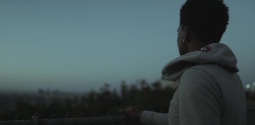 Someone adjusts a woman's hoodie as she stands at a fence overlooking a view