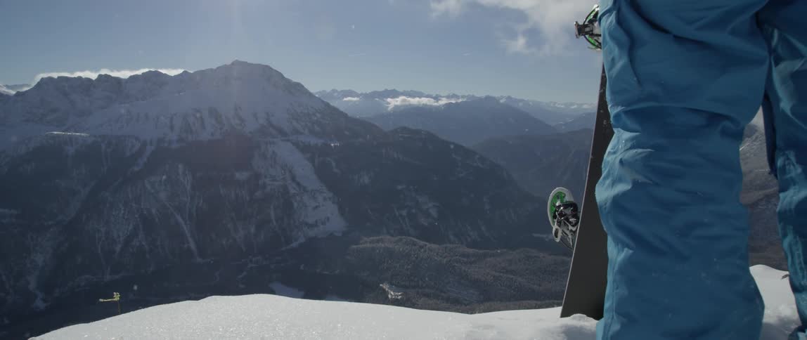 A man with a snowboard stands on a mountain peak taking in the view