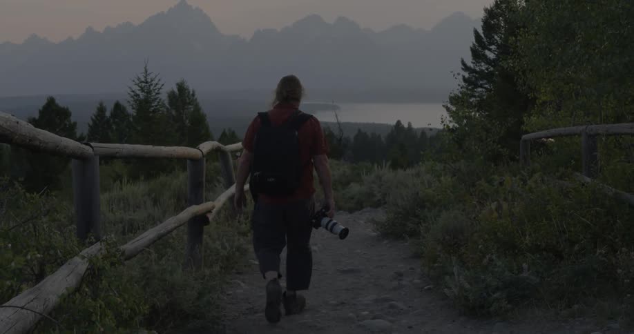 A person holding a camera walks along a path surrounded by grass