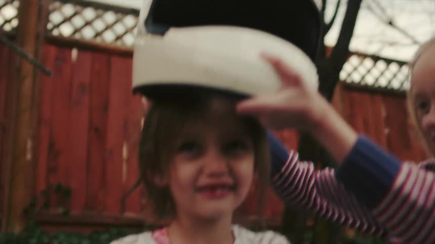 A girl places a helmet on another girl as they play in a backyard