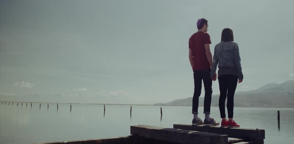 A man and woman hold hands as they stand on a pier looking at mountains