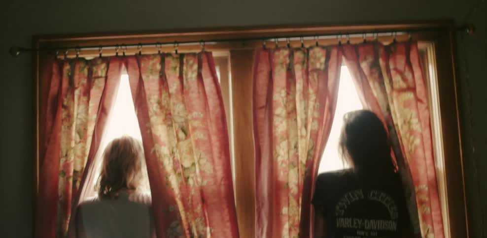 Two women stand between the curtains in windows and interact with each other