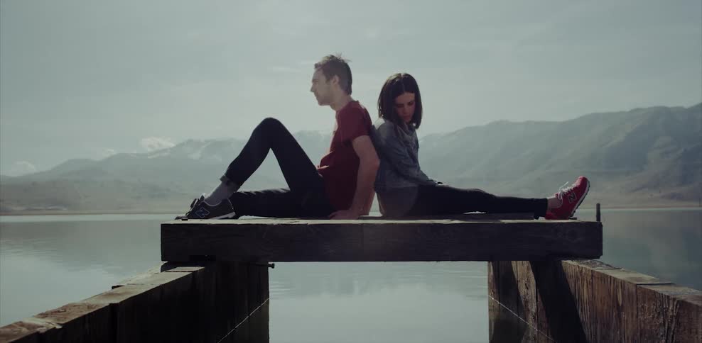 A man and woman sit back to back on a platform at a lake