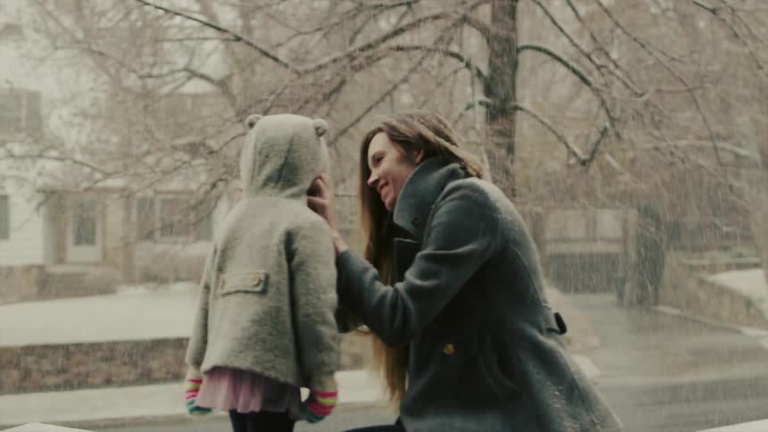 A woman pulls on a girl's coat hood while it snows