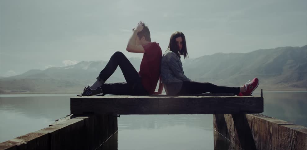 A man and woman sit back to back on a dock at a lake