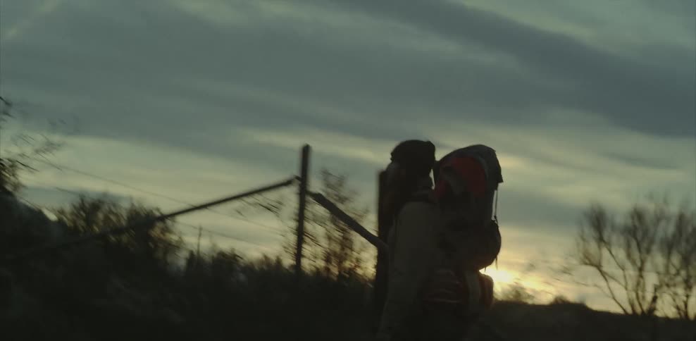 A man carrying a backpack is walking near a fence at dusk