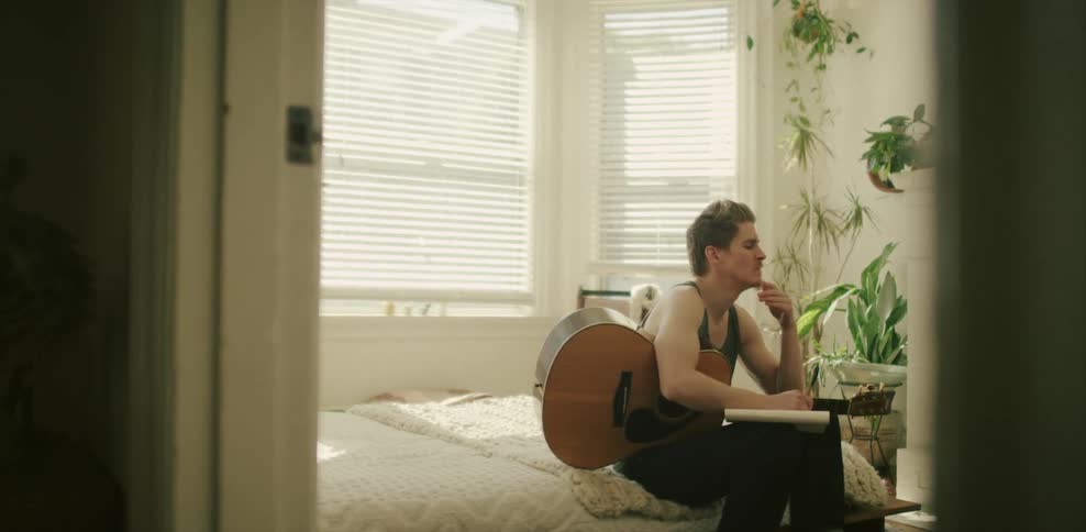 A guitarist holds a guitar and writes on paper on a bed