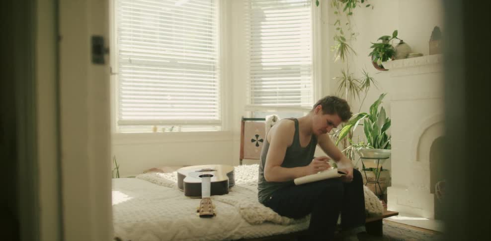 A man sits on a bed next to a guitar and writes in a notebook