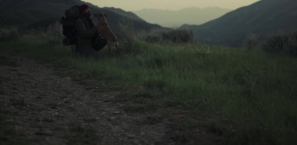 A man who is hiking is kneeling down petting a dog