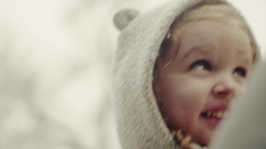 A woman holds a young girl and adjusts her coat hood while it snows