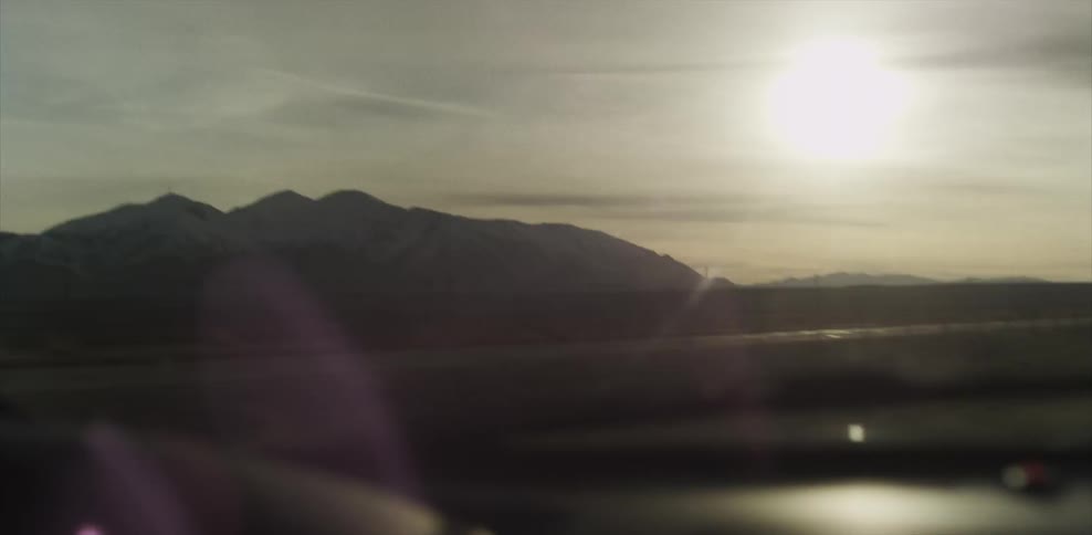A car drives on a rural road with the sun shining down on mountains
