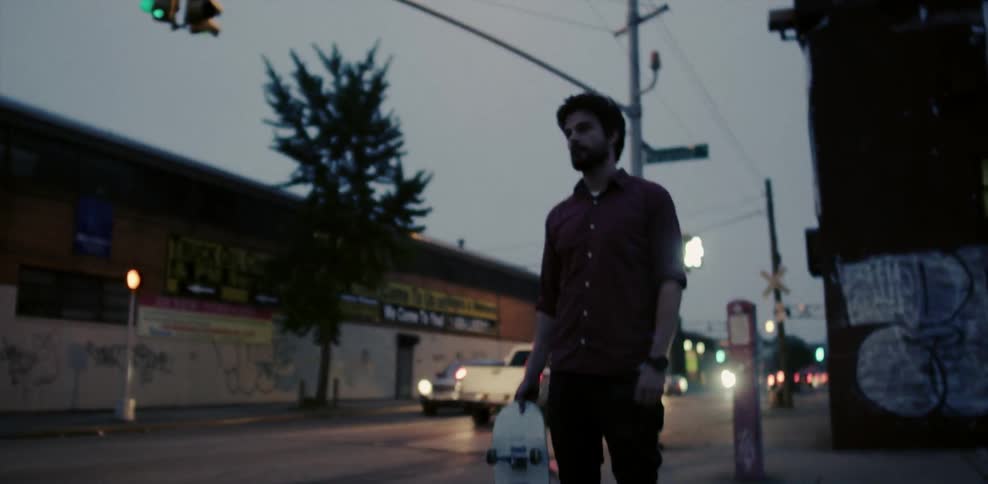 A man holding a skateboard stands in the middle of a crosswalk near a busy street at night