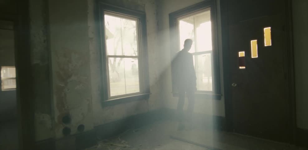 A man stands down the hall near the window in a dark home