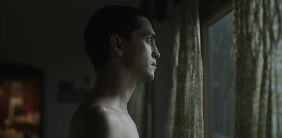 Shirtless man looks out a window