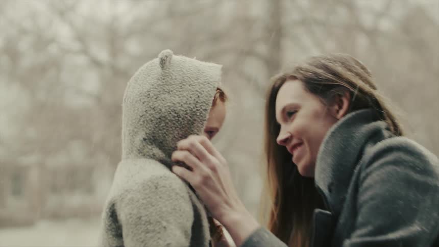 A woman pulls on a girl's coat hood while it snows outside