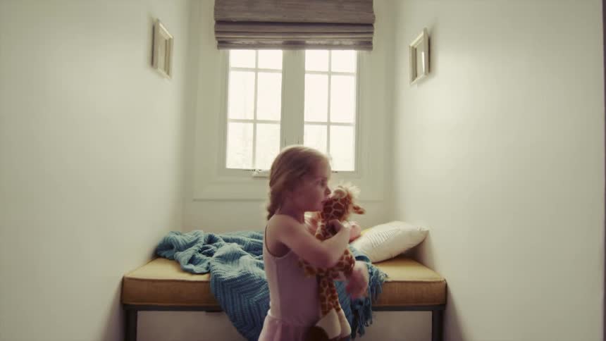 A little girl hugs a toy giraffe as she stands near a window during the day