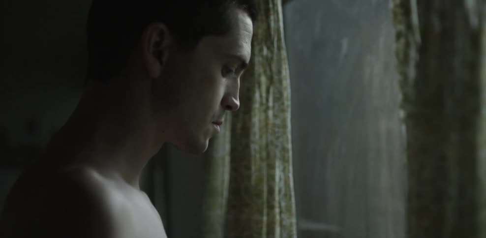A shirtless man stands next to a window