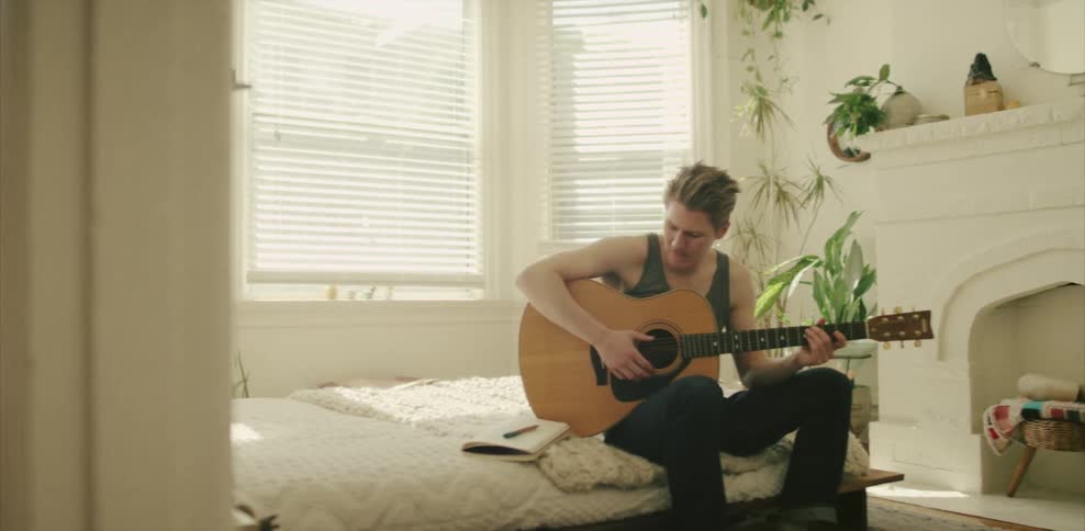 A man sitting on a bed strums a guitar and writes