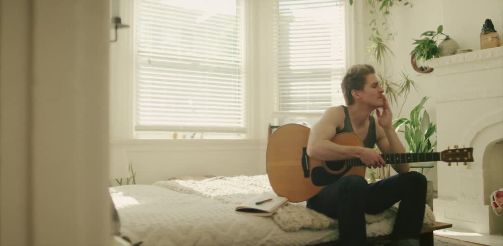 A guitarist holds a guitar and ponders while sitting on a bed