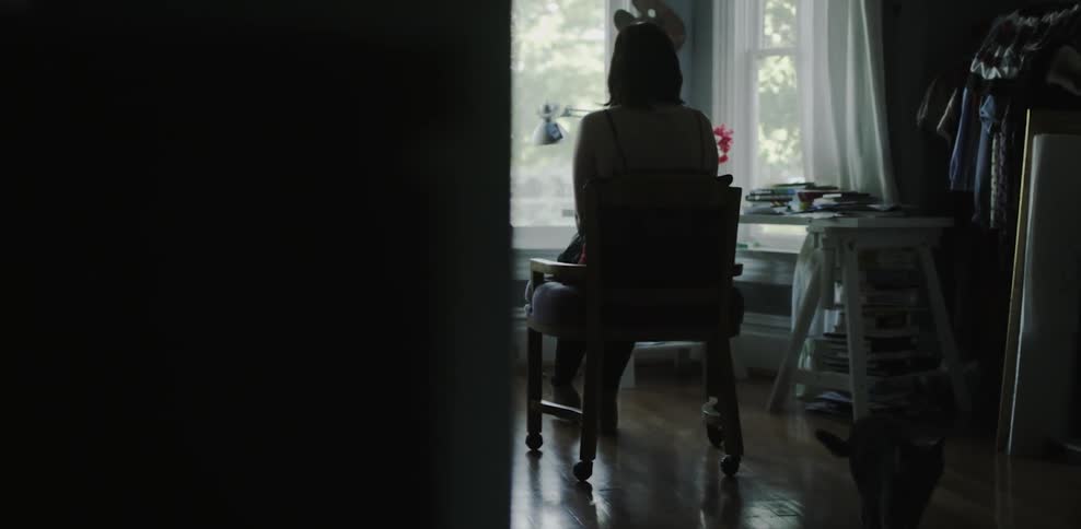 Woman sits on a chair looking out the window as a cat walks by