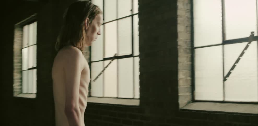 A thin man stands in an empty room and looks out the window