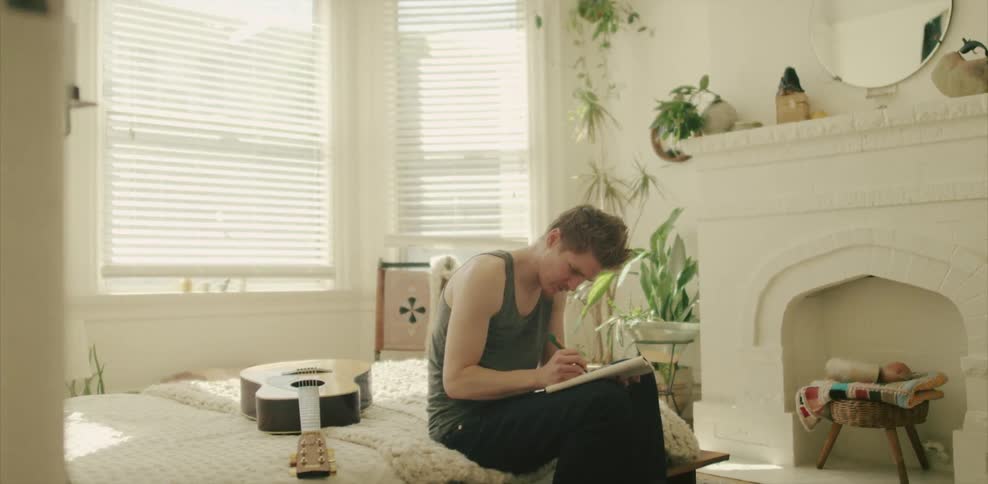 A man sitting on a bed with a guitar and writing on paper